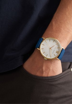 Zlata ura s teksturiranim belim številčnico, modrim usnjenim trakom, slim dizajnom in minimalističnimi urami. Nosijo jo na zapestju z sivo tkanino.