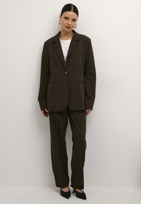 Mørkegrøn skræddersyet blazer med enkelt knaplukning, parret med matchende bukser, og en hvid t-shirt under. Sorte højhælede sko.