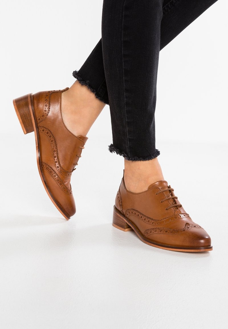 Chaussures brogues en cuir marron à lacets avec perforations décoratives, portées avec un jean noir à ourlet effiloché sur un sol clair.