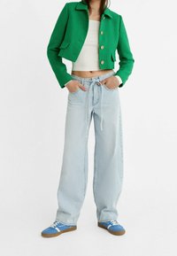 Kvinna klädd i en grön kort jacka, vit crop top, ljusblå vida jeans med snörning och blå sneakers med vita ränder.