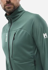 Grüne synthetische Jacke mit hohem Kragen, strukturierter Oberfläche und Reißverschlusstasche auf der Brust. Mit einem weißen Markenlogo am Ärmel.