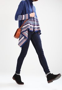 Person som går klädd i mörka jeans, svarta anklarstövlar, blå mönstrad cardigan, marinblå polotröja och bär en brun axelremsväska.