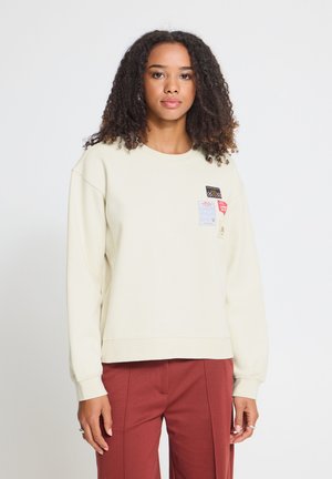 Frau mit lockigem Haar, die einen cremefarbenen Pullover mit Applikationen und rote Hose trägt, steht vor einem schlichten hellen Hintergrund.