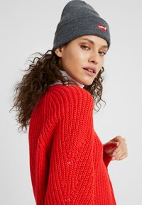 Rød strikket trøje med et struktureret mønster og en rund udskæring, parret med en grå ribbestrikket hue med et rødt Levi's-logo.