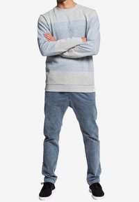 Sweatshirt gris clair et bleu à rayures horizontales, avec un logo. Associé à un pantalon bleu fuselé et des chaussures noires.