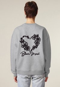 Sweatshirt grigio con un design floreale a forma di cuore e la scritta "Bloom Mood" ricamata in nero sul retro. Presenta una vestibilità rilassata e polsini a coste.