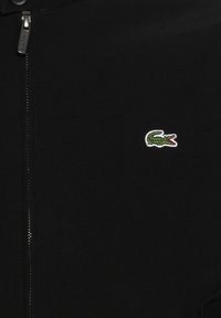 Veste noire avec fermeture éclair à l'avant montrant un petit logo crocodile vert brodé sur la poitrine.