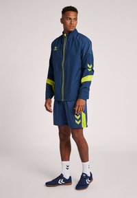 Marinblå idrottsjacka med gula accenter, dragkedja fram och logo; matchas med marinblå shorts med grönt zigzag-mönster och matchande skor.