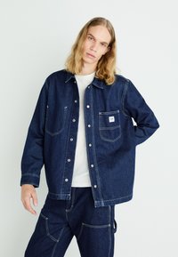 Giacca in denim blu scuro con maniche lunghe, bottoni a pressione, doppie tasche sul petto e dettagli di cucitura bianca, abbinata a una t-shirt bianca.