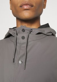 Grå vattentät jacka med huva, med tryckknappsstängning, justerbara dragsnören och subtila silverdetaljer. Slät, matt textur.