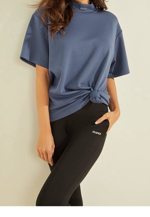 Femme portant un haut bleu à manches courtes noué à la taille et un legging noir Guess, main dans la poche, sur un fond uni.