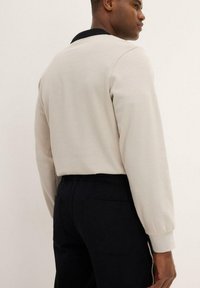 Felpa di un beige chiaro con colletto nero, caratterizzata da maniche lunghe e una vestibilità comoda. Abbinata a pantaloni scuri dotati di tasche laterali.