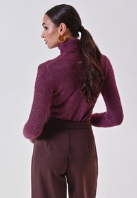 Donna con una lunga coda di cavallo scura, che indossa orecchini a cerchio dorati, un maglione a collo alto viola e pantaloni marroni a vita alta, di spalle.