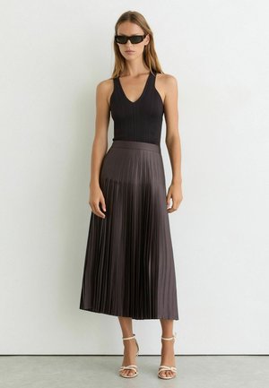 Mujer con top negro sin mangas, falda midi plisada marrón oscuro, gafas de sol negras y sandalias blancas de tacón alto con tiras, de pie contra una pared blanca.