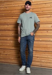 Camiseta de manga corta verde claro con logotipo, jeans azules y zapatillas blancas. El modelo tiene tatuajes y está de pie contra una pared de madera.