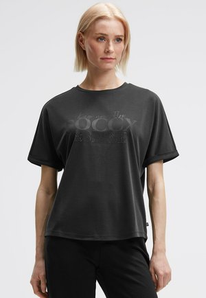 Frau mit kurzem blonden Haar trägt ein lockeres schwarzes T-Shirt mit aufgerollten Ärmeln und dezentem Text "Behalte deinen Fluss & strahle" auf der Vorderseite.