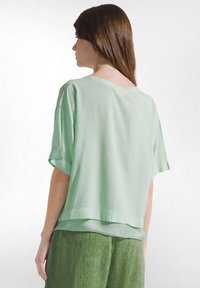 Kvinde med langt brunt hår iført løs lysegrøn top med korte ærmer og mørkegrønne bukser, set bagfra mod en hvid baggrund.