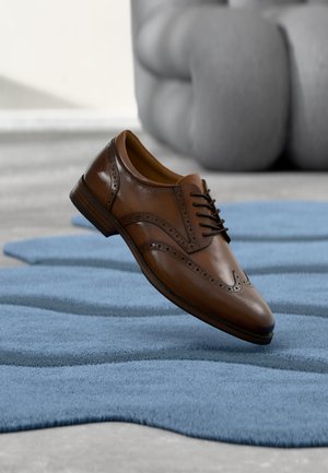 Scarpa elegante brogue in pelle marrone fluttuante sopra un tappeto blu strutturato con una sedia imbottita grigia sullo sfondo sfocato.