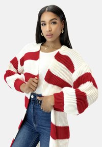 Cárdigan de punto grueso con rayas rojas y blancas, de frente abierto, mangas largas, textura acanalada, superpuesto a una camiseta blanca y combinado con jeans azules.