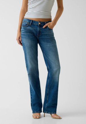 Frau trägt blaue Jeans mit geradem Bein und der Hand in der Tasche, kombiniert mit einem weiß-schwarzen gestreiften ärmellosen Top und goldenen High-Heel-Sandalen.