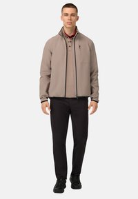 Veste zippée marron clair avec un col montant, finitions foncées au niveau du zip, et une poche poitrine. Portée par-dessus un polo sombre et un pantalon noir, avec des chaussures noires.