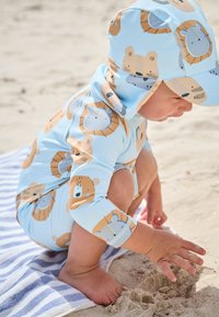 Light blue swim outfit with bear patterns, long sleeves, and a matching hat. Worn by a child crouching on sand, interacting with the surface.