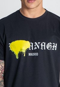 Čierne bavlnené tričko s nápismi "MALANAGH" a "MADRID" s živým dizajnom žltých farieb rozliatych na tkanine, čo vytvára kontrastný efekt.