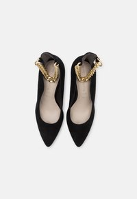 Ballerines pointues en daim noir avec des brides épaisses en chaîne dorée aux chevilles, vues de dessus sur un fond clair uni.