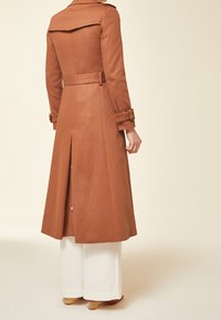 Hnědý trench coat se strukturovaným designem, velkým límcem a páskem v pase. Obsahuje zadní rozparek a knoflíkové detaily podél lemování. Pod ním jsou bílé kalhoty.