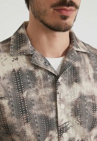 Chemise à manches courtes avec un motif en nuances de beige et de marron. Elle possède un col et une fermeture à boutons à l'avant, fabriquée en tissu léger.