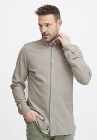 Chemise à manches longues beige clair avec une texture douce. Présente un col boutonné et un petit accent rayé près de l'ourlet.