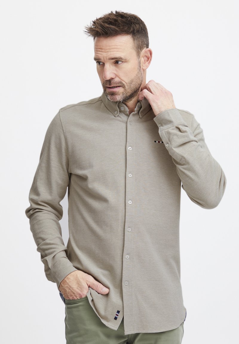 Chemise à manches longues beige clair avec une texture douce. Présente un col boutonné et un petit accent rayé près de l'ourlet.