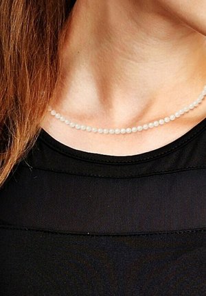 Collier en perles blanches, espacées de manière homogène, porté autour du cou. Déposé sur un haut noir texturé, mettant en avant le design minimal de l'accessoire.