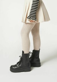 Czarne skórzane buty wojskowe z masywną podeszwą, sznurowane i z zamkiem błyskawicznym z boku, zestawione z jasnymi, prążkowanymi rajstopami i plisowaną, jasną spódnicą.