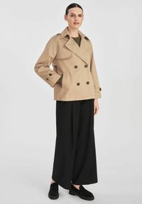 Beżowy dwurzędowy płaszcz trench z naramiennikami, dużymi guzikami i kieszeniami bocznymi, zestawiony z szerokimi czarnymi spodniami i czarnymi butami.
