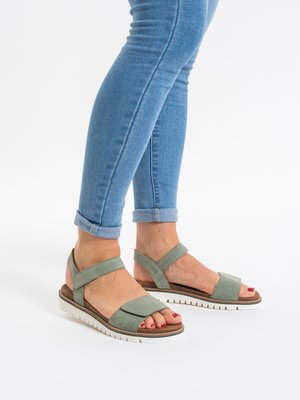 Pieds en jean bleu clair retroussé portant des sandales vertes à bout ouvert avec des semelles blanches sur fond blanc.