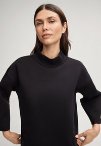 Zwarte pullover top met een hoge, geribde hals, relaxte pasvorm en zachte textuur. Heeft zijzakken en brede 3/4-lange mouwen.