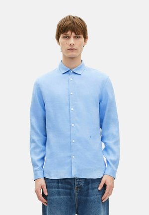 Jeune homme aux cheveux bruns courts, portant une chemise bleue claire boutonnée et un jean bleu, debout devant un fond blanc uni.