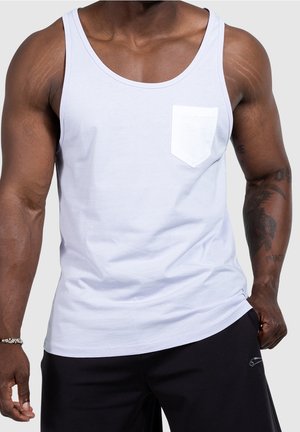 Muskularna osoba ubrana w biały tank top z kieszonką i czarne spodnie, stojąca na jednolitym tle.