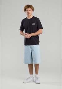 Black cotton T-shirt with a small white graphic on the chest, paired with light blue denim shorts and white sneakers.