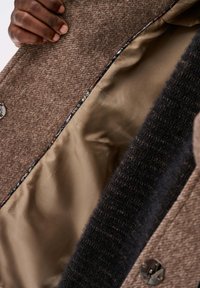 Bruin getextureerde wollen jas met een gladde beige satijnen voering, voorzien van decoratieve binnennaad en metalen knoopdetails.