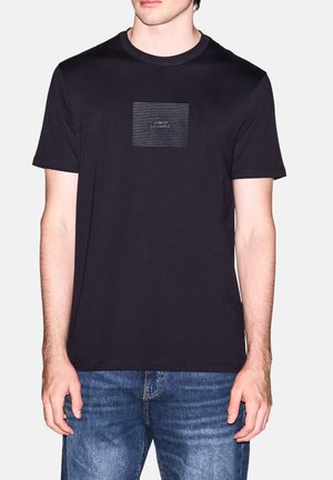 Mężczyzna noszący czarną koszulkę Armani Exchange z białymi poziomymi liniami i logo na piersi, zestawioną z niebieskimi jeansami.