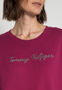 Sudadera fucsia con cuello redondo acanalado y el texto bordado en plata "Tommy Hilfiger" en el pecho. Textura suave y diseño casual.