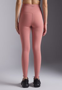 Høyhalsede rosa treningstights i et glatt, elastisk stoff, med flate sømmer og en logo i midjen, kombinert med svarte joggesko.