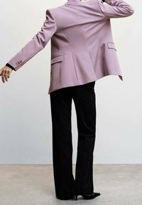 Blazer lilas sur mesure avec épaules structurées et deux poches arrière, associé à un pantalon noir à jambes larges et des talons pointus.