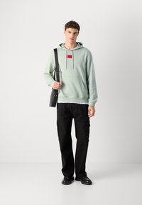 Joven con sudadera verde claro con parche de logo rojo, pantalones cargo negros, zapatos negros y llevando una bolsa de hombro negra, de pie contra un fondo blanco.