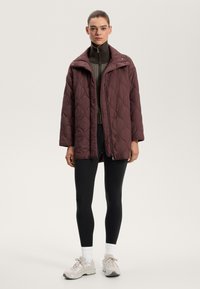 Gequilted bordeaux jacket met een brede kraag, drukknoopsluitingen en een relaxte pasvorm, gedragen over een aansluitend bruin topje en zwarte leggings, gecombineerd met lichte schoenen.