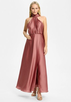 Femme blonde portant une robe de soirée en satin rose à col licou avec une fente, des talons dorés à brides et des boucles d’oreilles dorées imposantes sur fond blanc.