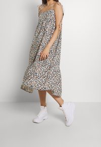 Blumiges Midi-Kleid mit gerafftem Oberteil, verstellbaren Bindegurten und einem rüschengesäumten Saum. Das Muster zeigt kleine rosa und blaue Blumen; kombiniert mit weißen Stiefeln.
