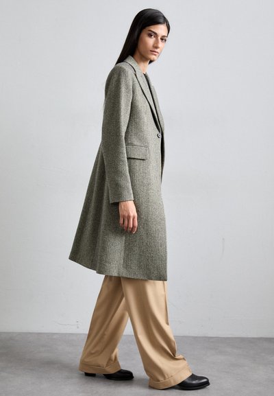 Herringbone patterned long coat in shades of grey, featuring notch lapels, a single button closure, and side pockets, paired with wide beige trousers.